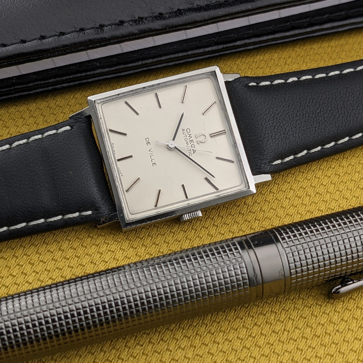 Omega De Ville Automatik 161.014 diagonal neben strukturiertem silbernem Stift und schwarzem Gehäuse, mit hellem Zifferblatt und schwarzem Armband sichtbar.