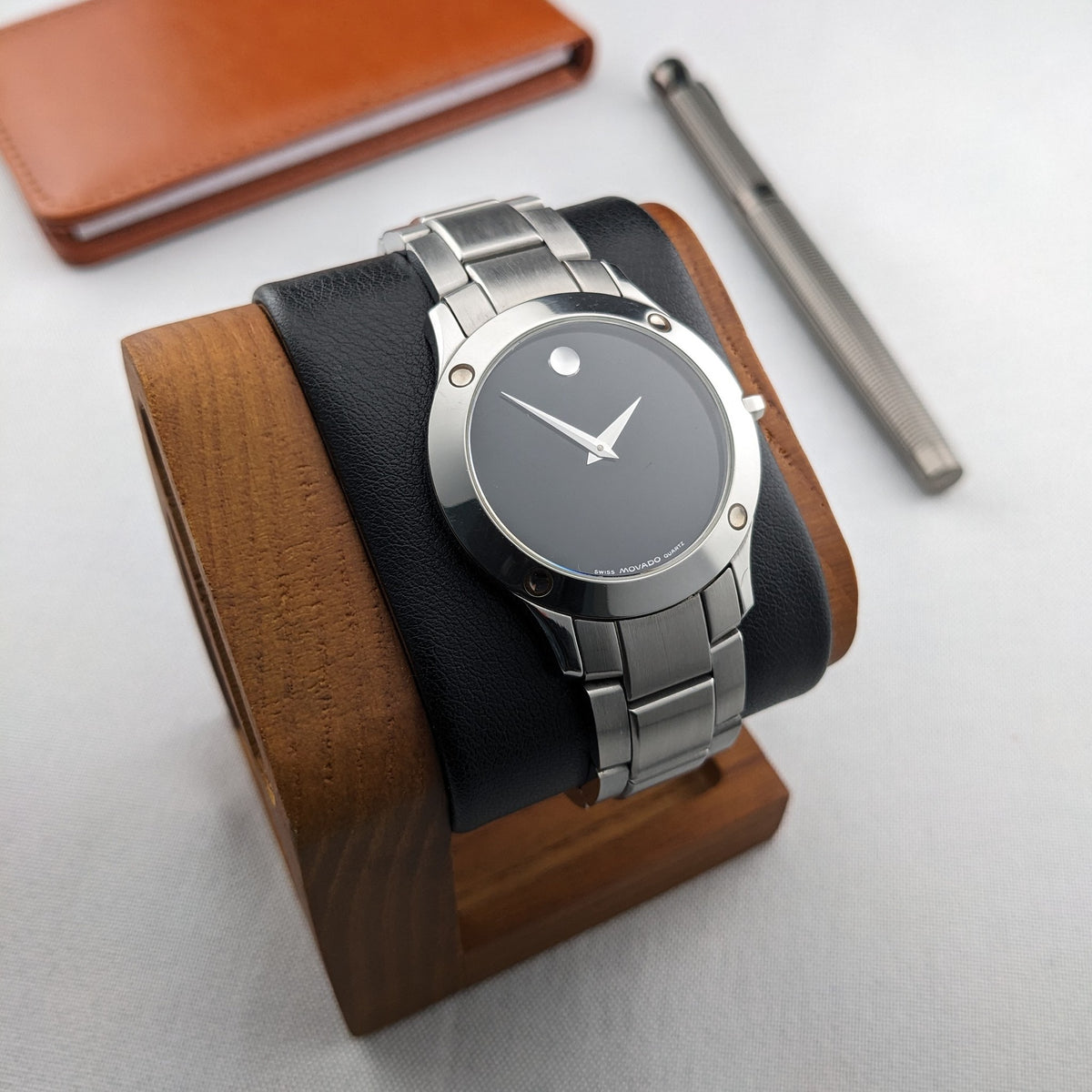 Montre Movado montée sur un support d'exposition noir et bois, avec un étui brun et un stylo foncé en arrière-plan.