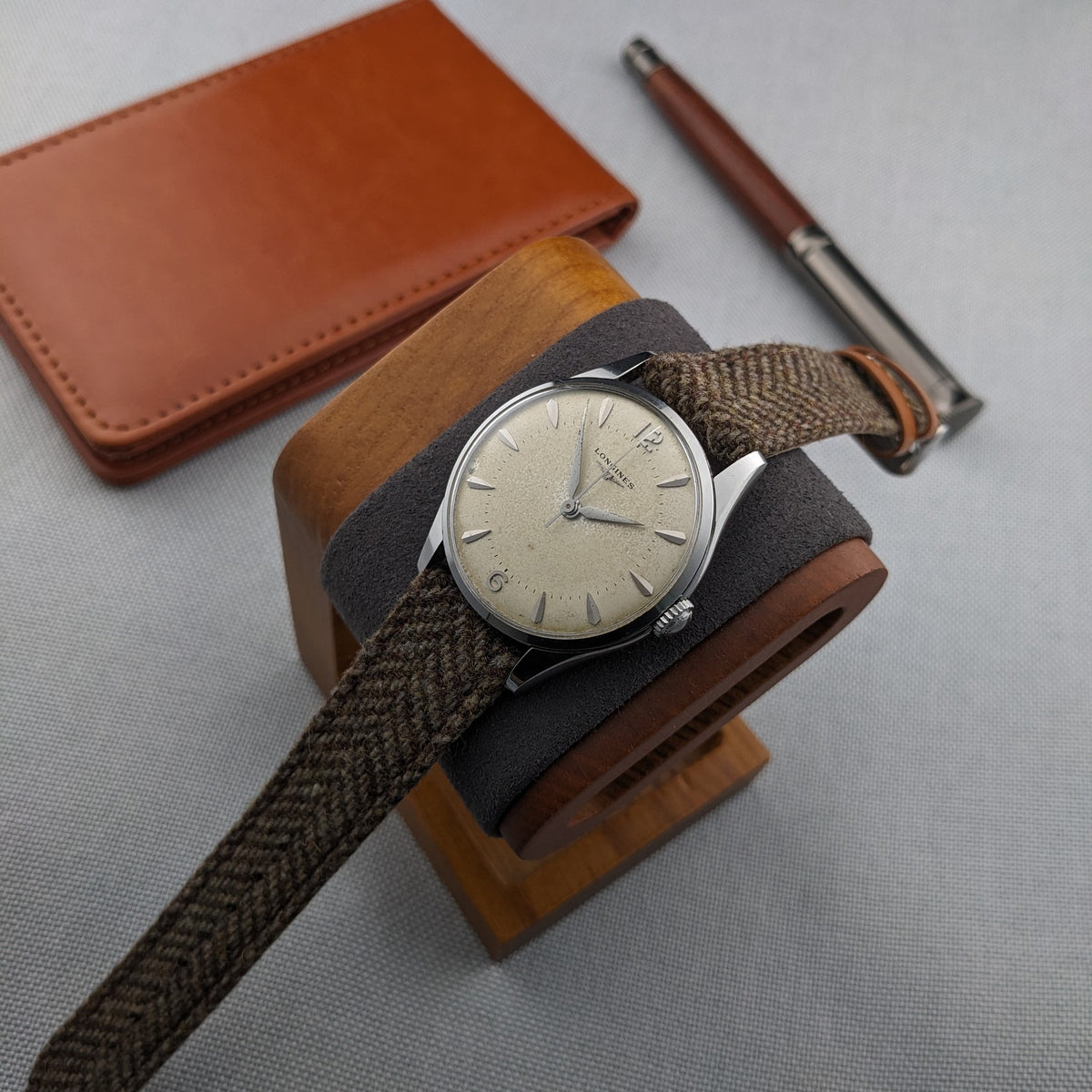Longines Classic reference 8135-5 draped over a wooden watch stand, with a leather wallet and pen arranged in the background.