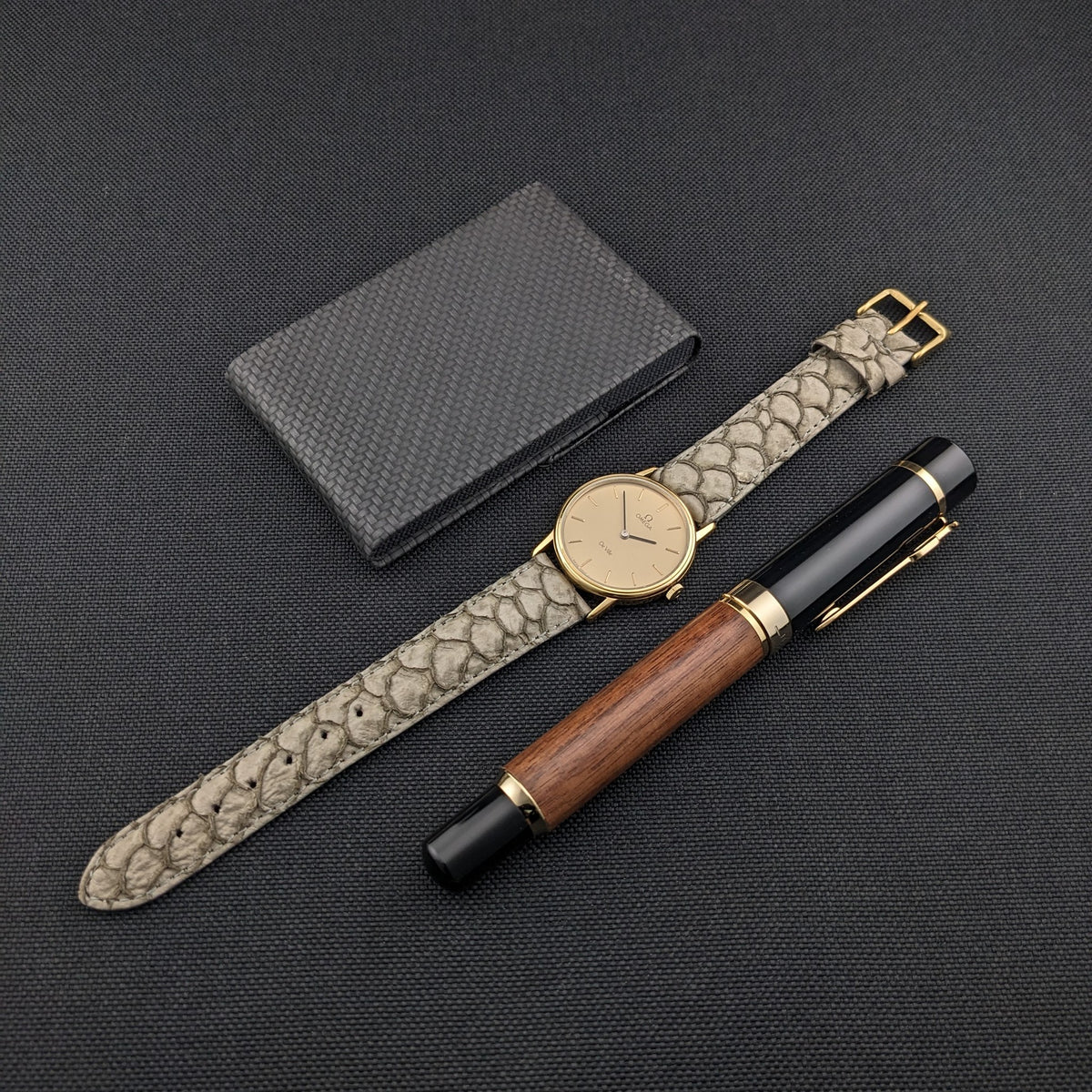 Omega De Ville reference 111 0156, gray strap extended beside a dark notebook and wood-and-black pen on a black surface.