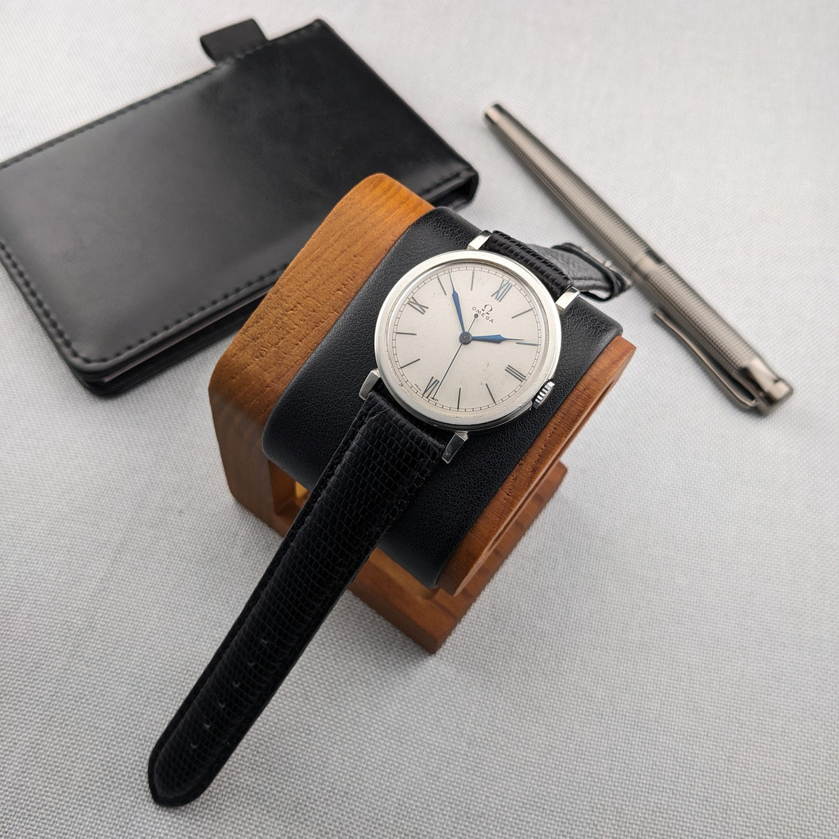 Omega watch displayed on a wood-and-black cushion with the strap extending forward, beside a black wallet and silver pen.