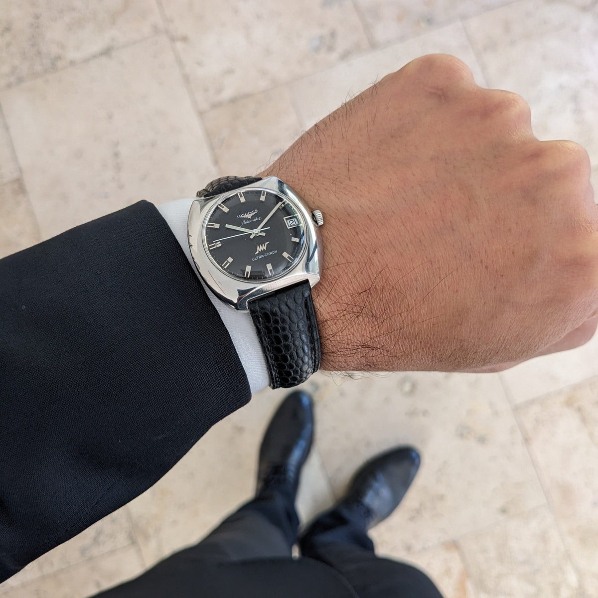 Longines Ultrachron reference 8429 pictured on a wrist above a tiled floor, with dark dial, date display and black leather strap visible.