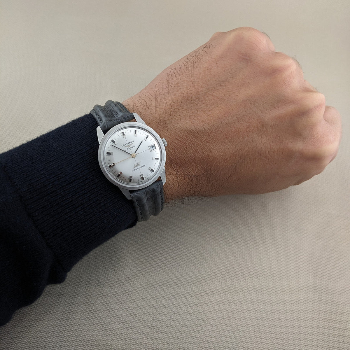 Longines Ultra-Chron 7827 worn on a wrist, showing the silver dial and black strap against a dark sleeve and neutral background.