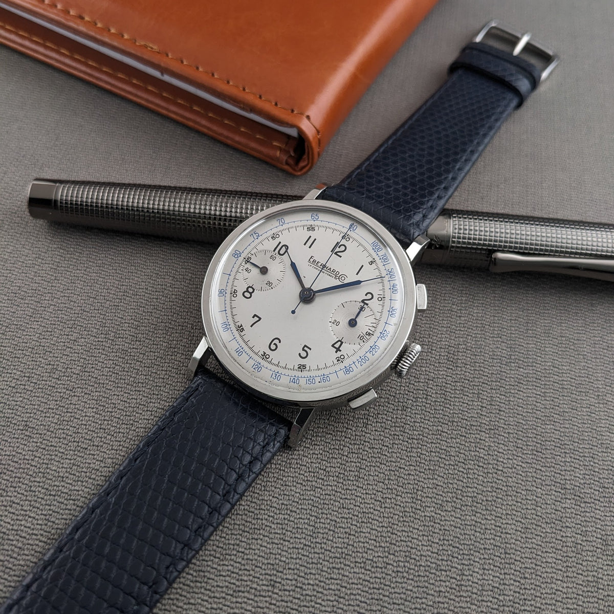 Eberhard & Co.. Pre-Extra Fort is centered on a blue strap over a textured pen, with a brown wallet in the background.