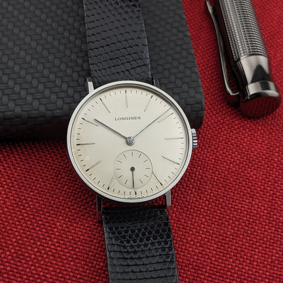 Straight-on Longines Calatrava reference 20757 dial composition, placed over a black notebook and beside a silver pen on red fabric.