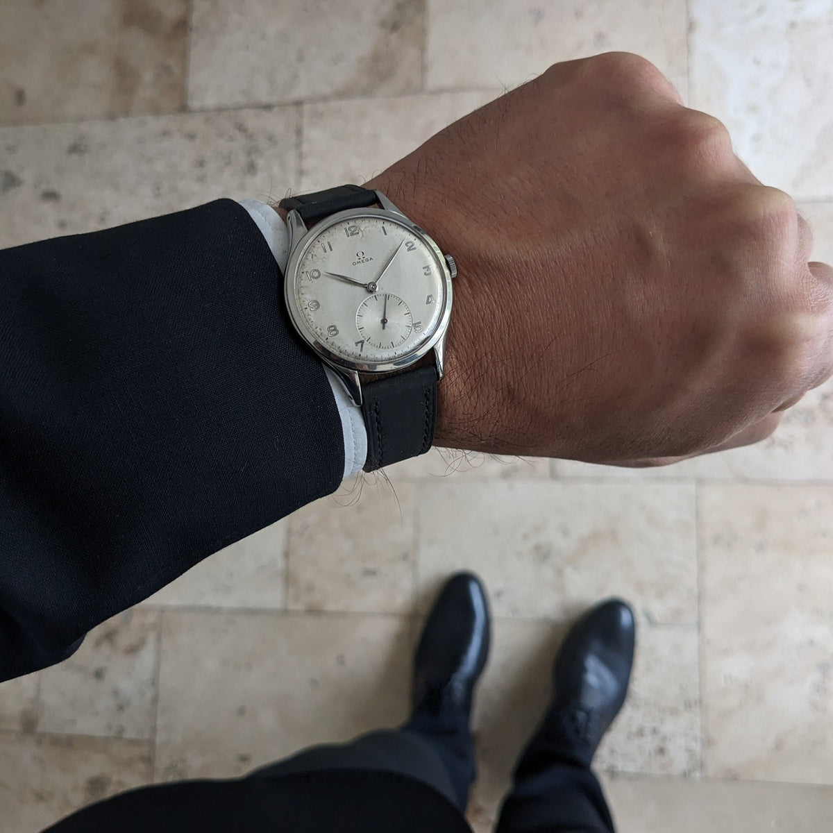 Wrist view of the Omega Jumbo reference 2505-5 worn with a dark suit, photographed above a tiled floor with black shoes visible.