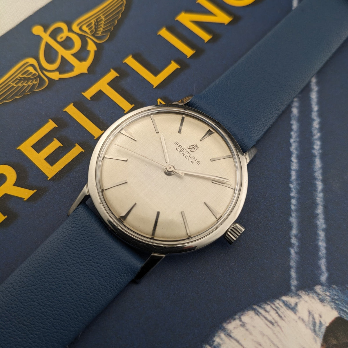 Breitling Automatic watch displayed on a dark blue Breitling-covered book, with the cream dial and blue strap prominent.