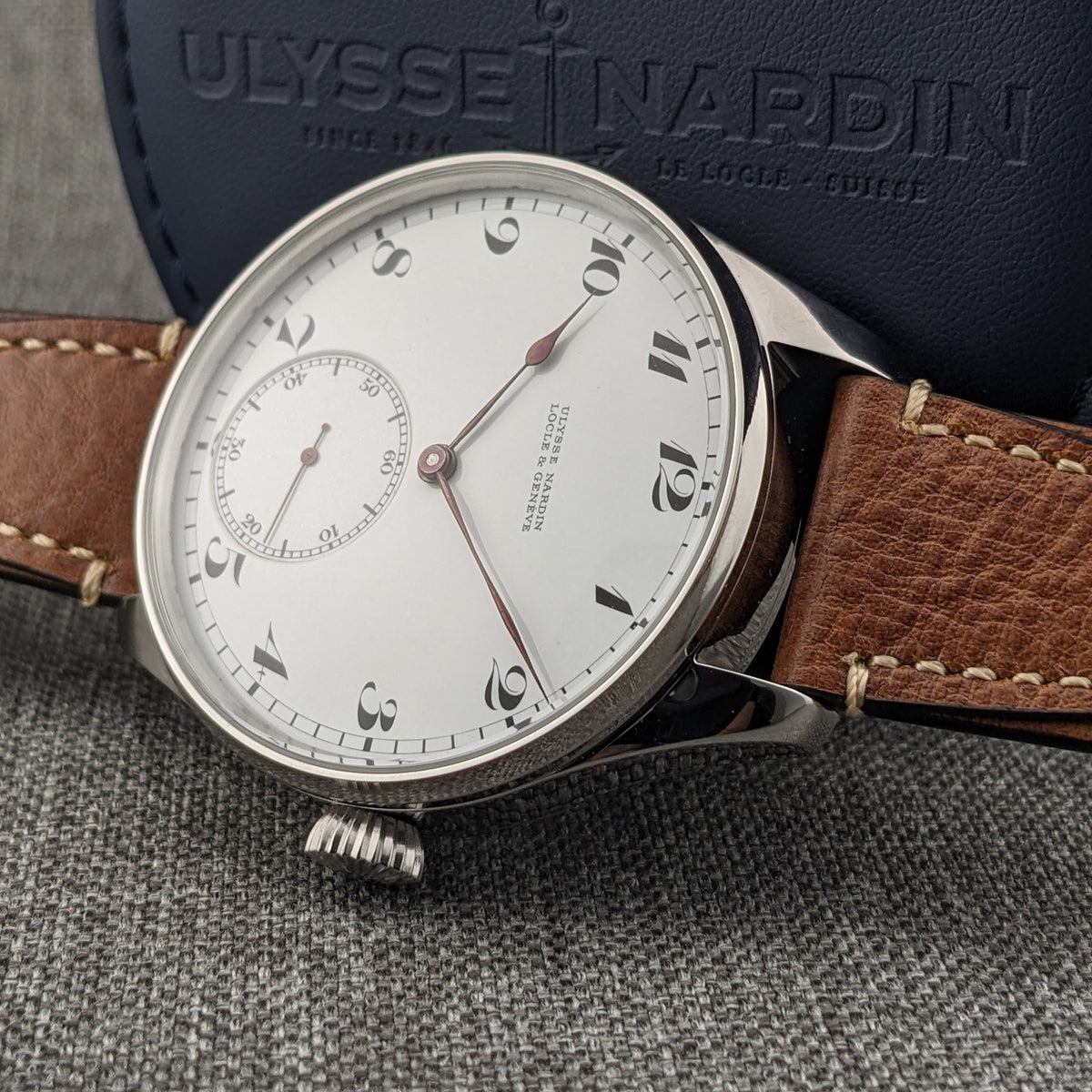 Ulysse Nardin कलाई घड़ी नीले ब्रांडेड केस पर कोणीय, सफेद अंक डायल, छोटे सेकंड रजिस्टर और रिड्ज़ क्राउन के साथ।