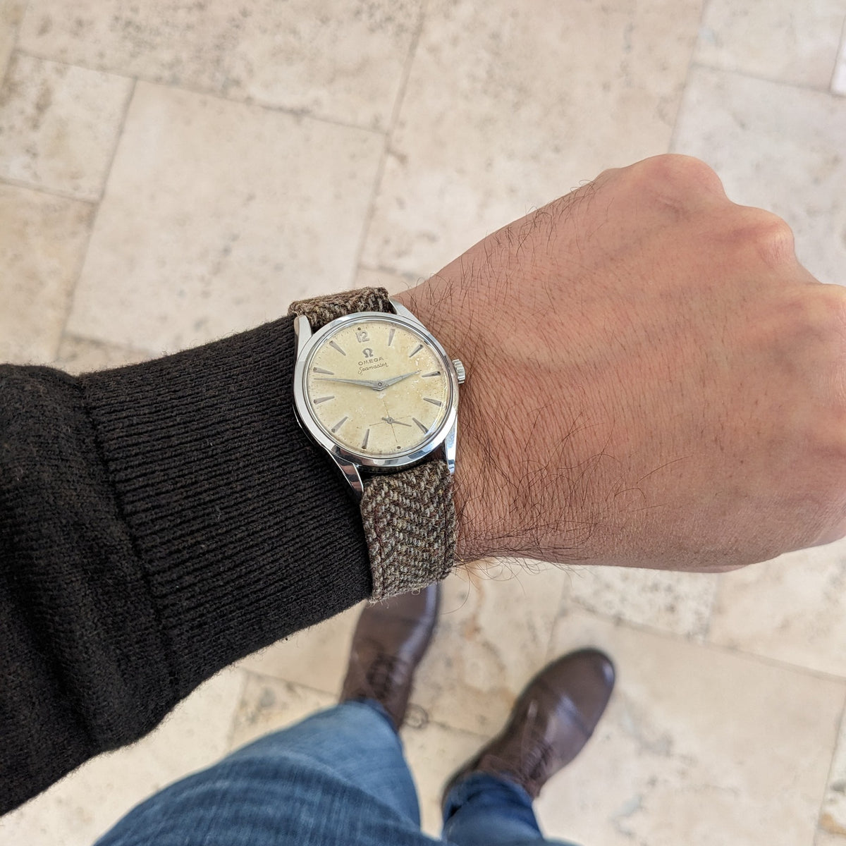 Omega Seamaster reference 29374 shown on a wrist from a wider view, with cream dial, woven strap, black sleeve and tiled floor.