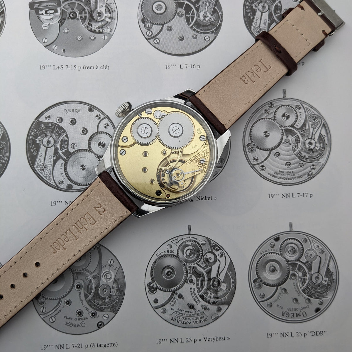 Orologio Omega visto dal lato del movimento su una pagina di meccanismi illustrati, con ingranaggi dorati e cinturino chiaro visibili.