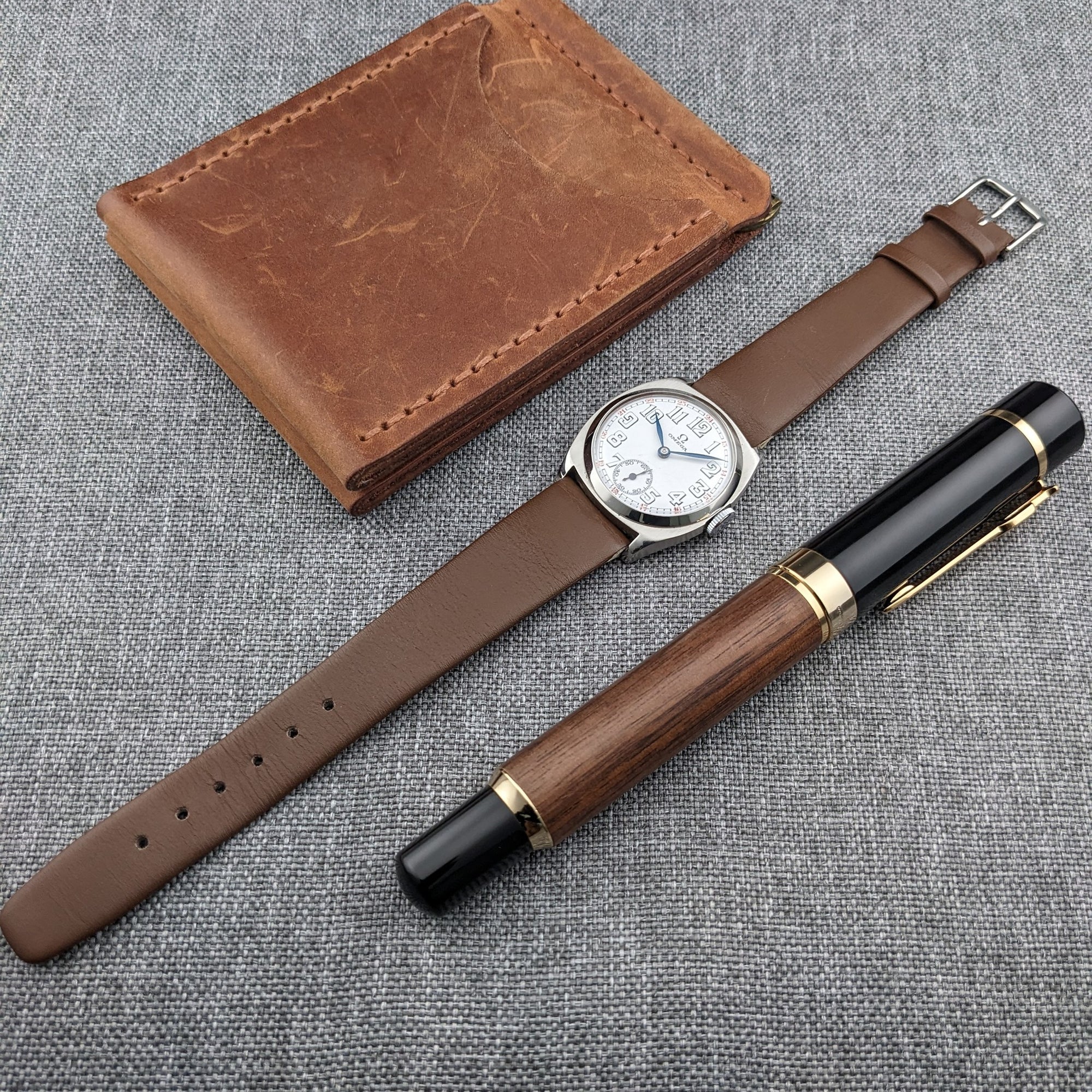 Omega watch, extended brown leather strap, worn brown wallet, and black-and-wood pen arranged together on gray woven fabric.