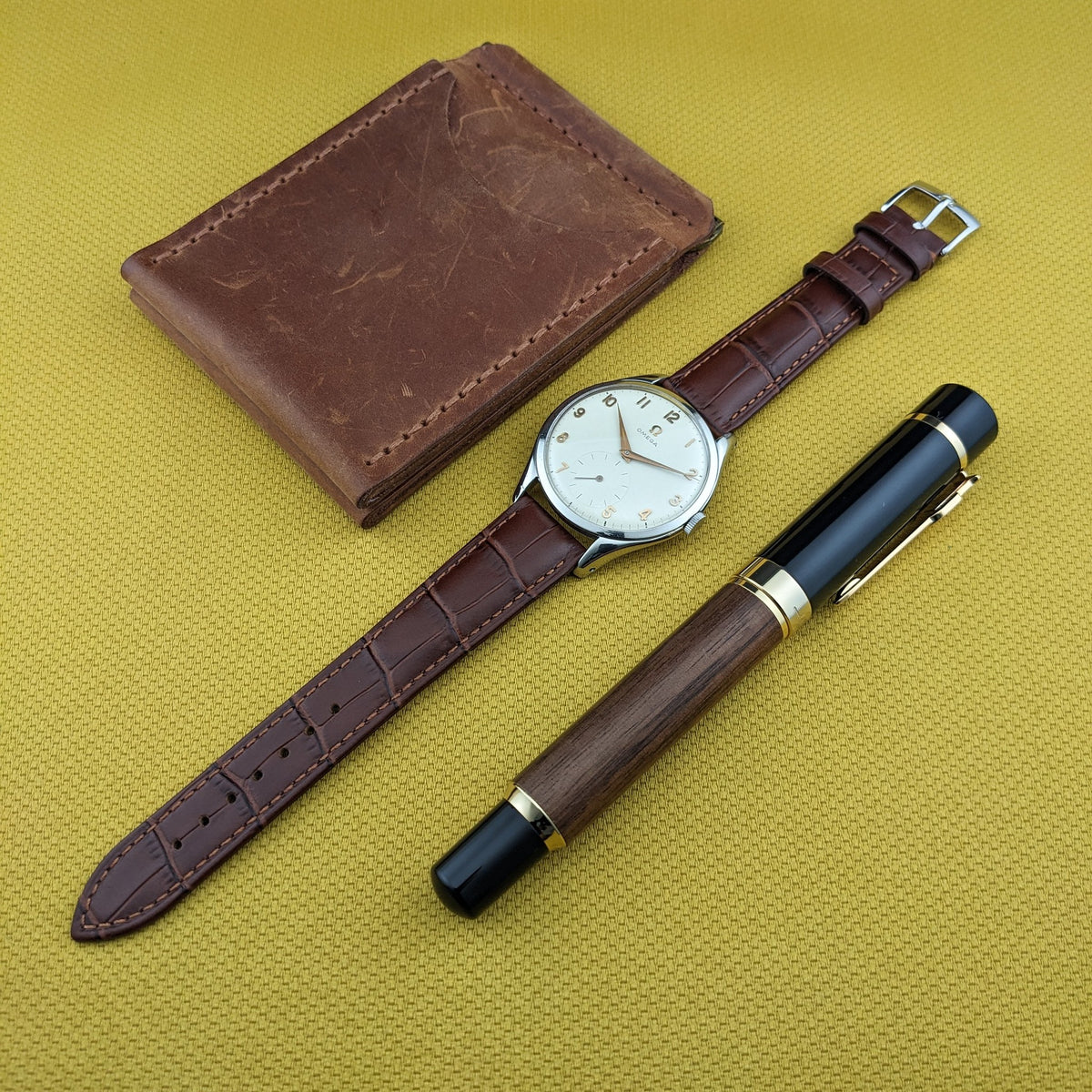 Omega Jumbo Referenz 2505-10, braunes Portemonnaie und Holzstift auf goldener Oberfläche arrangiert.
