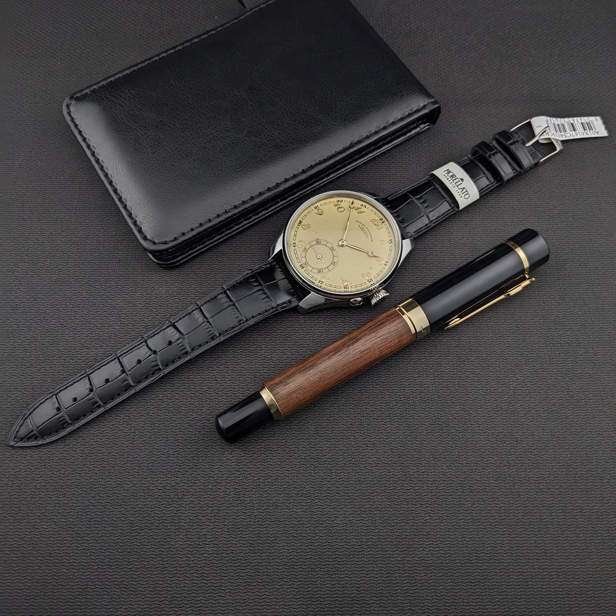 Gesamtansicht der Deutsche Präzisions-Uhrenfabrik Glashütte Uhr, schwarze Geldbörse und Holz-und-schwarzer Stift auf dunklem Stoff.
