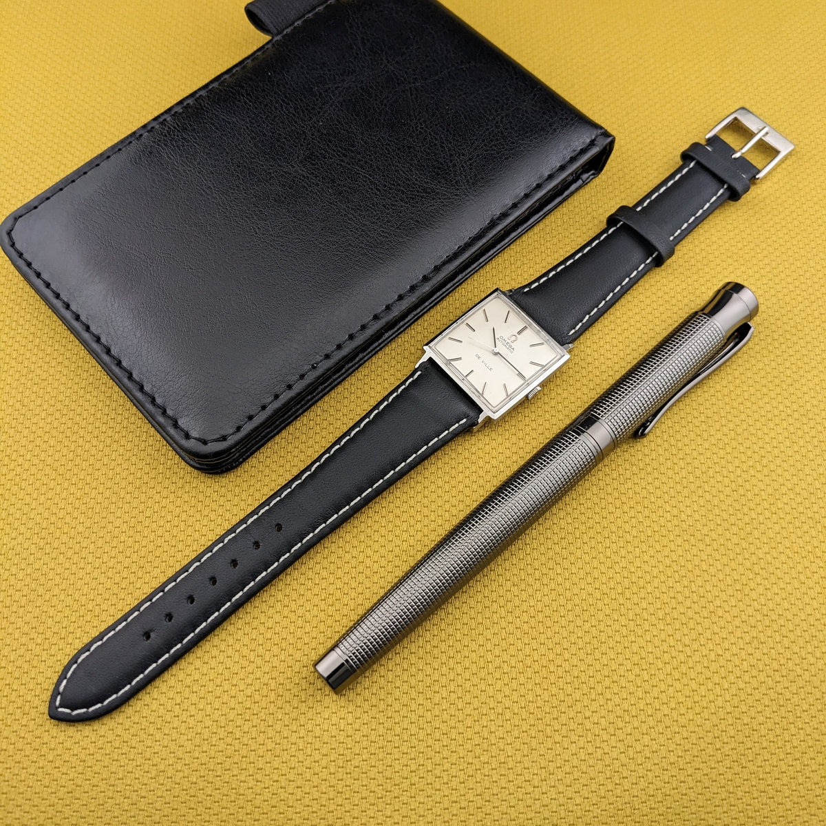 Omega De Ville Automatik 161.014 mit ausgezogenem schwarzen Armband, schwarzem Gehäuse und silbernem Stift auf senffarbenem Untergrund.