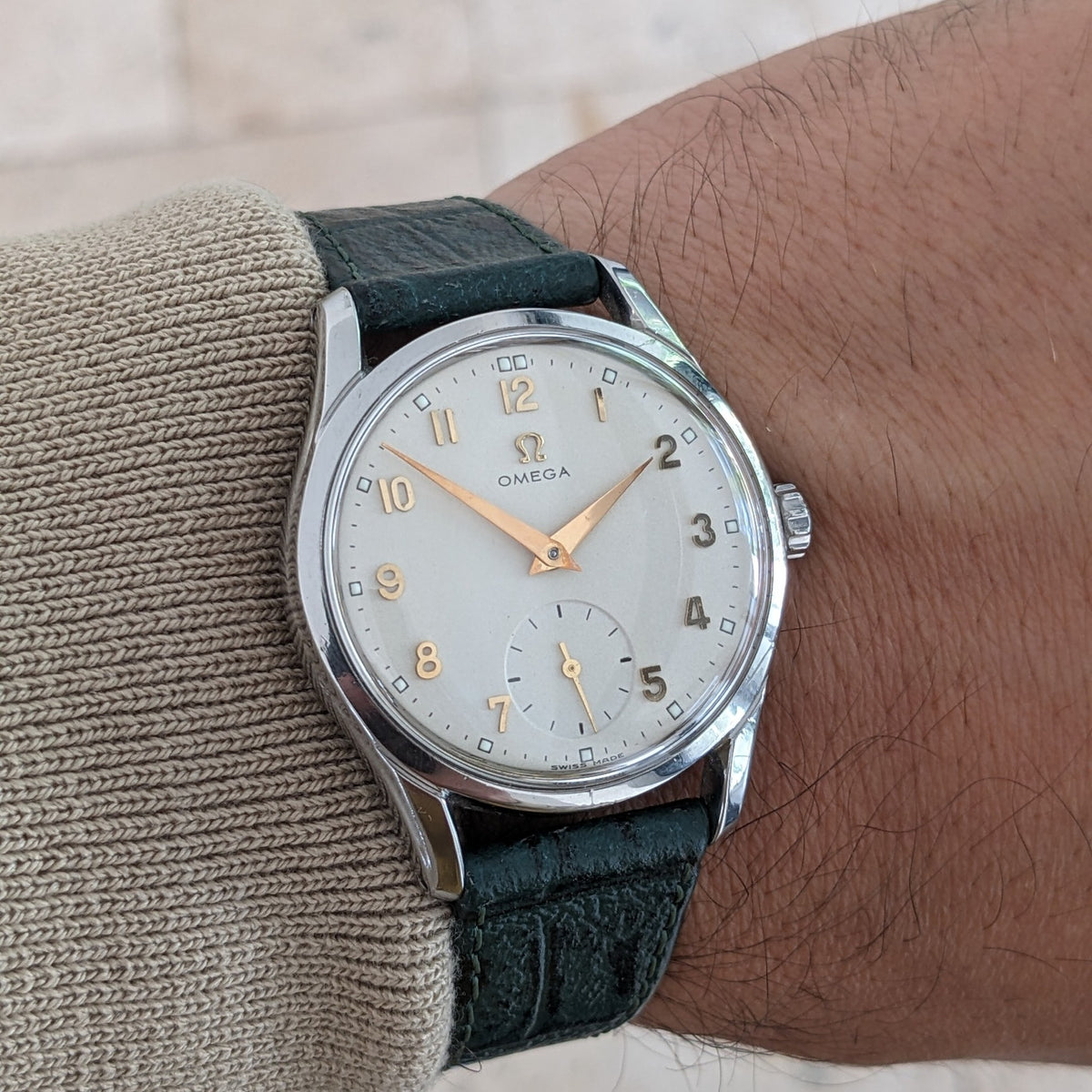 Vista polso ravvicinata dell'OMEGA Classic 2639-11 con quadrante argentato, numeri dorati, cinturino verde, manica beige e pavimento piastrellato.