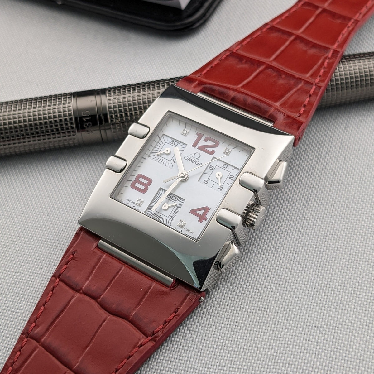 Omega Constellation Quadra 1841.73.31 on a red strap placed beside a textured metal pen and a partially visible black case.
