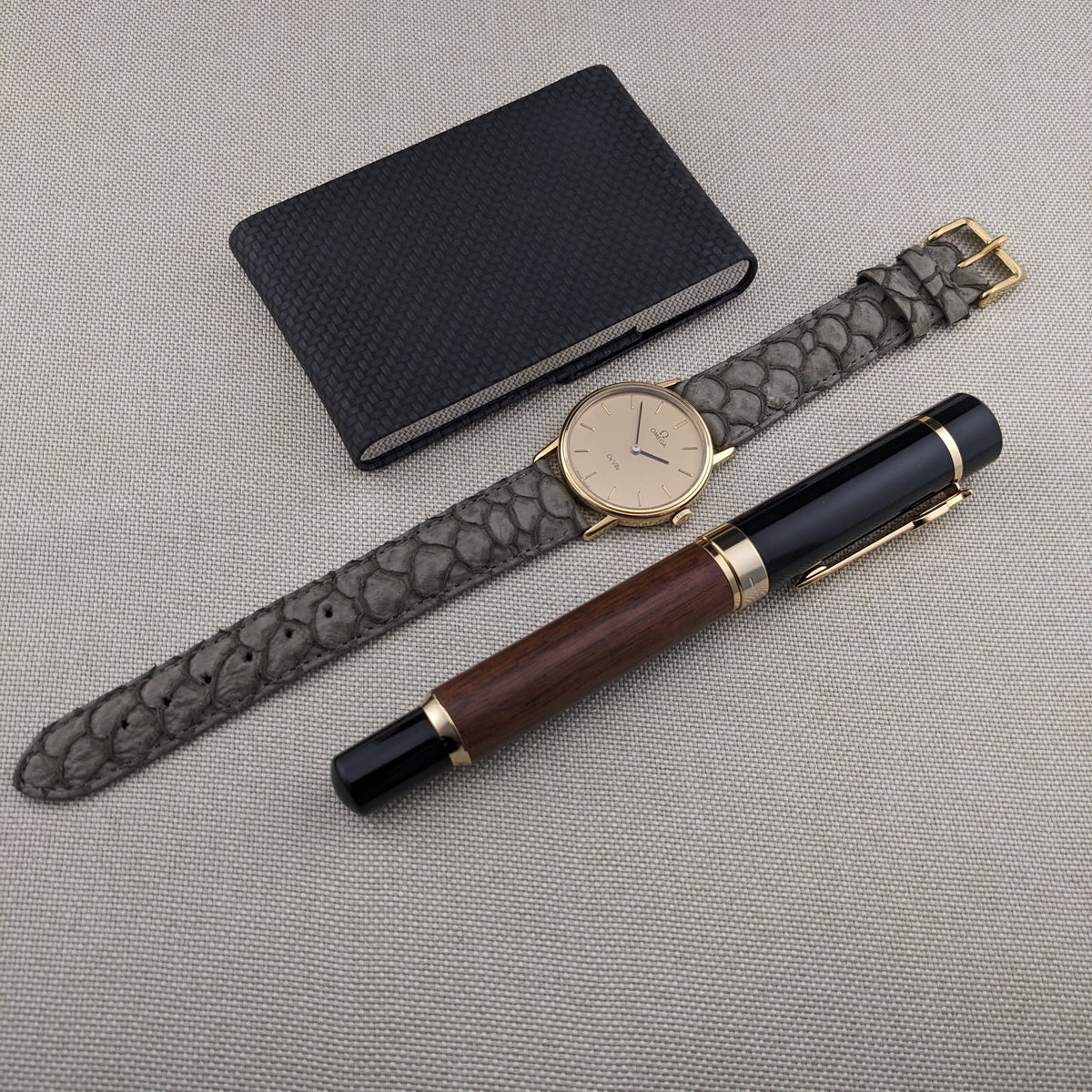 Omega De Ville reference 111 0156 arranged with its gray strap, dark notebook, and wood-and-black pen on a light woven background.