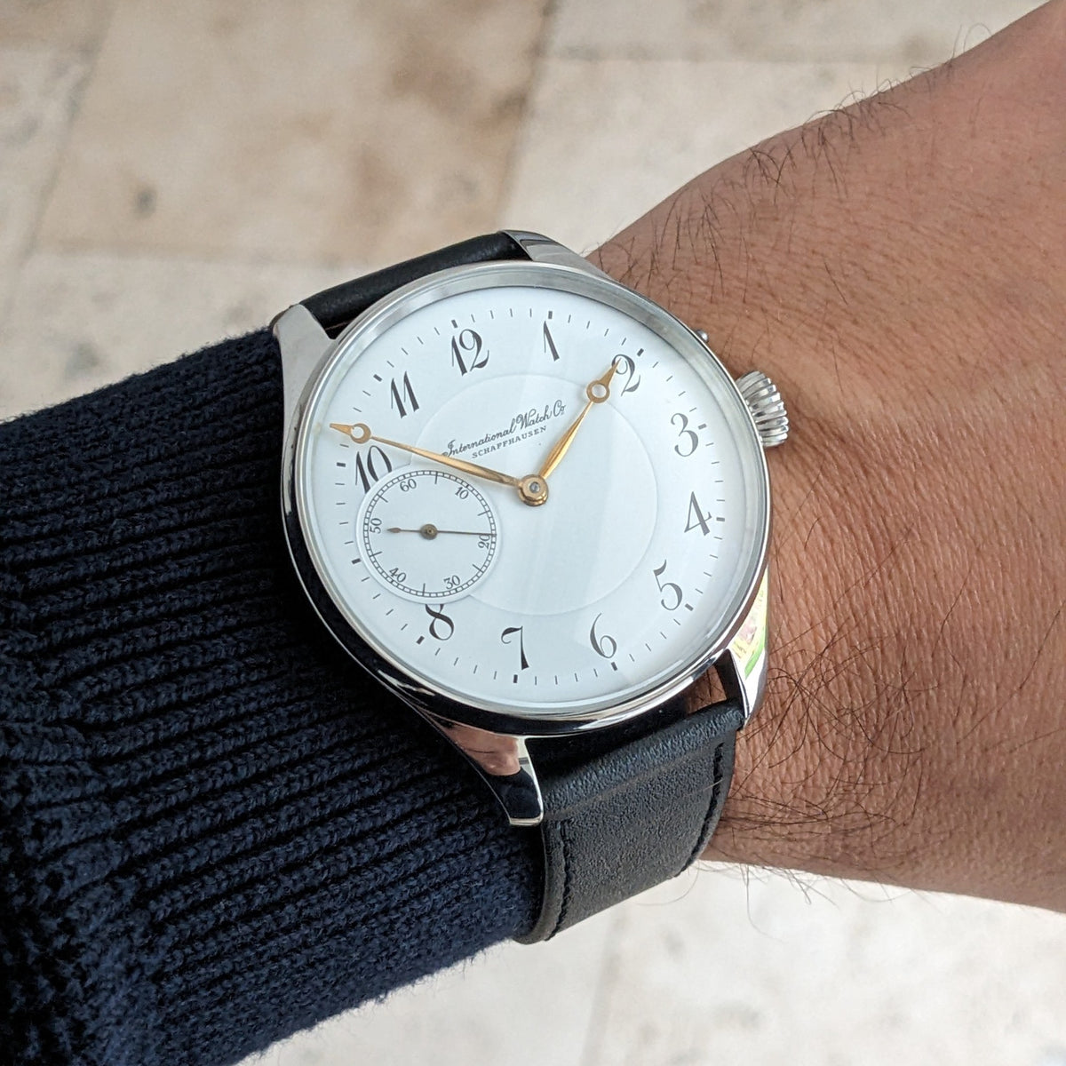 IWC Schaffhausen कलाई घड़ी पहने हुए, सफेद अंक, सुनहरे हाथ, छोटे सेकंड्स डायल और काले चमड़े की पट्टी के साथ।