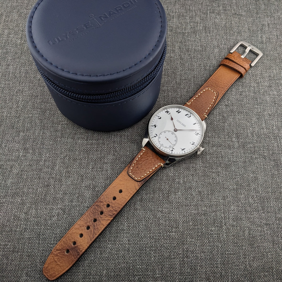 Ulysse Nardin कलाई घड़ी एक गोल नीले ज़िप केस के बगल में रखी हुई, जिसमें भूरा चमड़े का पट्टा ग्रे कपड़े की सतह पर विस्तारित।
