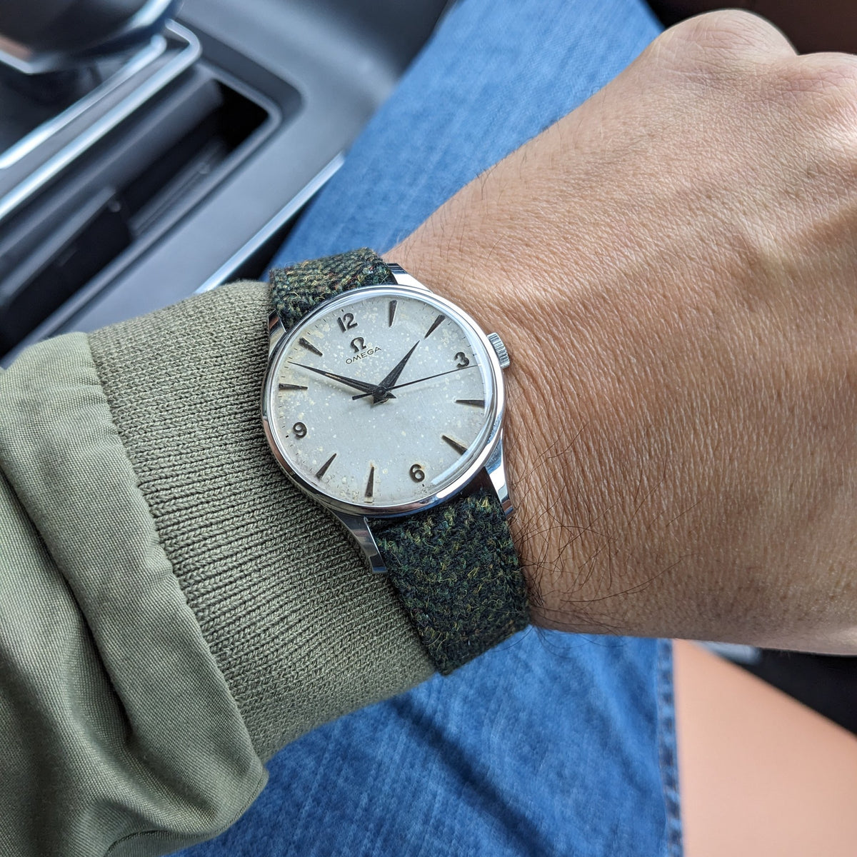 Am Handgelenk getragene Omega Classic Referenz 2810-2 SC neben einem Fahrzeuginnenraum fotografiert, mit sichtbarem hellem Zifferblatt und grünem Armband.