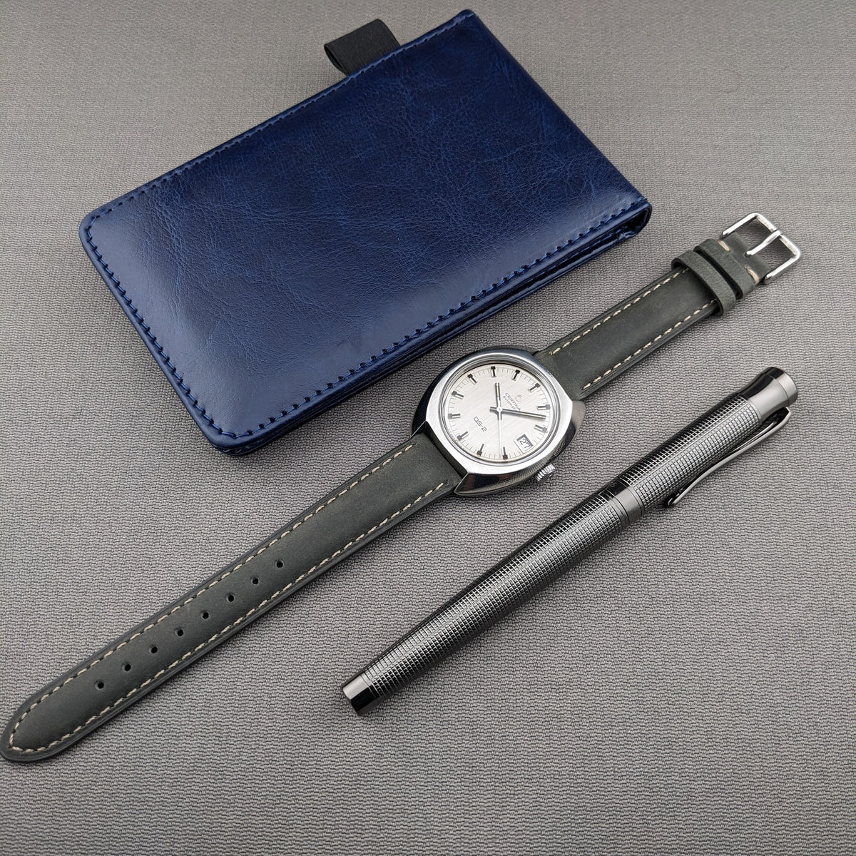 Full arrangement featuring the Certina DS-2 Automatic reference 5801 300, extended dark strap, blue wallet, and textured metal pen.
