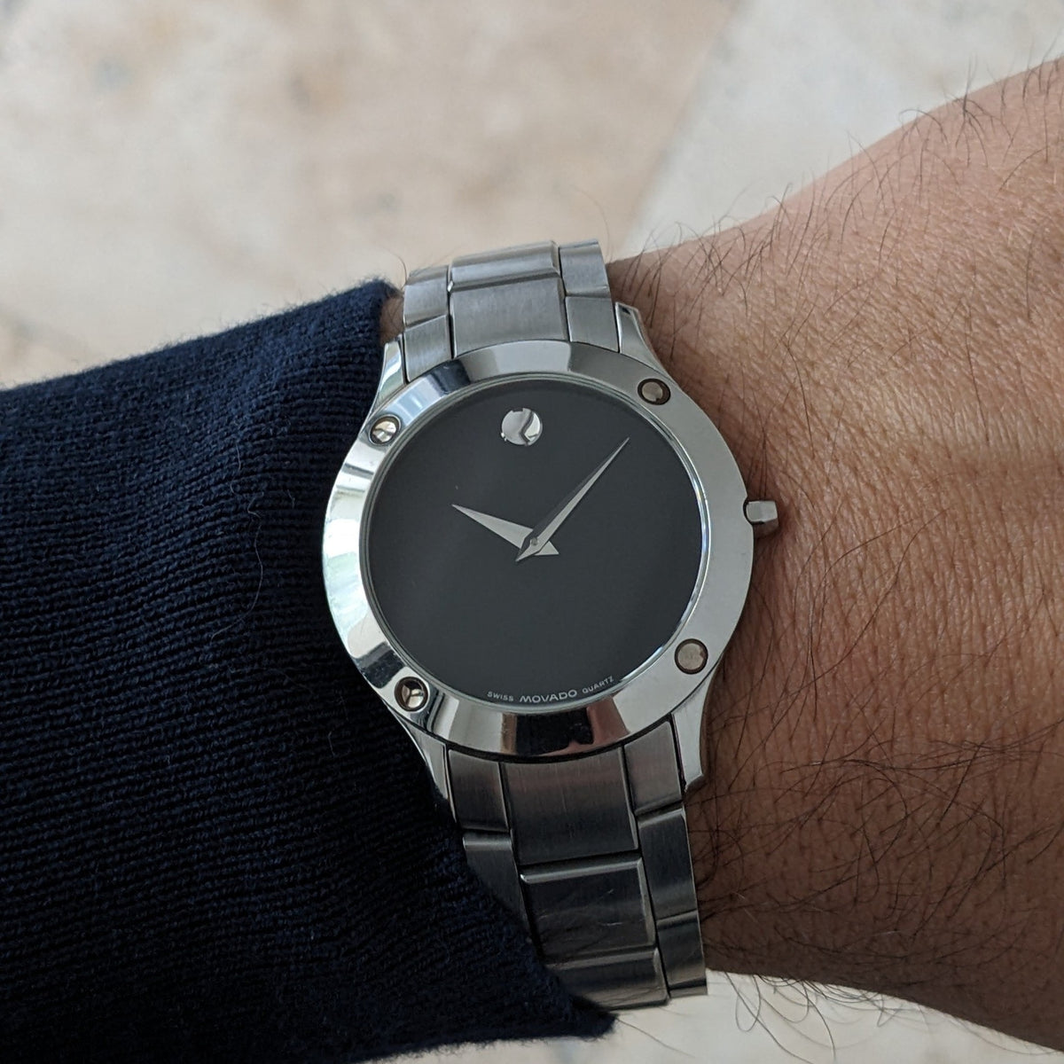 Montre Movado portée au poignet, montrant le cadran noir, aiguilles argentées, repère circulaire, attaches de lunette et bracelet en acier.