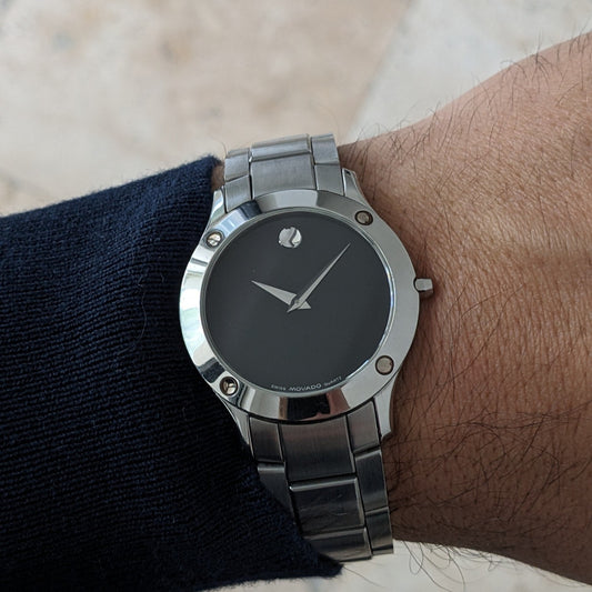 Movado Uhr am Handgelenk getragen, zeigt das schwarze Zifferblatt, silberne Zeiger, kreisförmigen Marker, Lünettenbefestigungen und Stahlarmband.