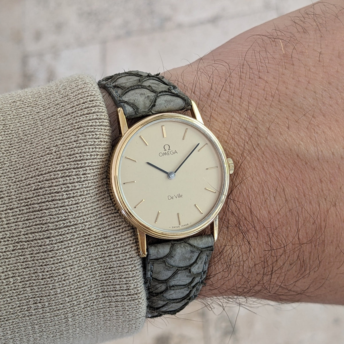 Omega De Ville reference 111 0156 worn on a wrist, showing the cream dial, gold-tone case, and gray textured strap.