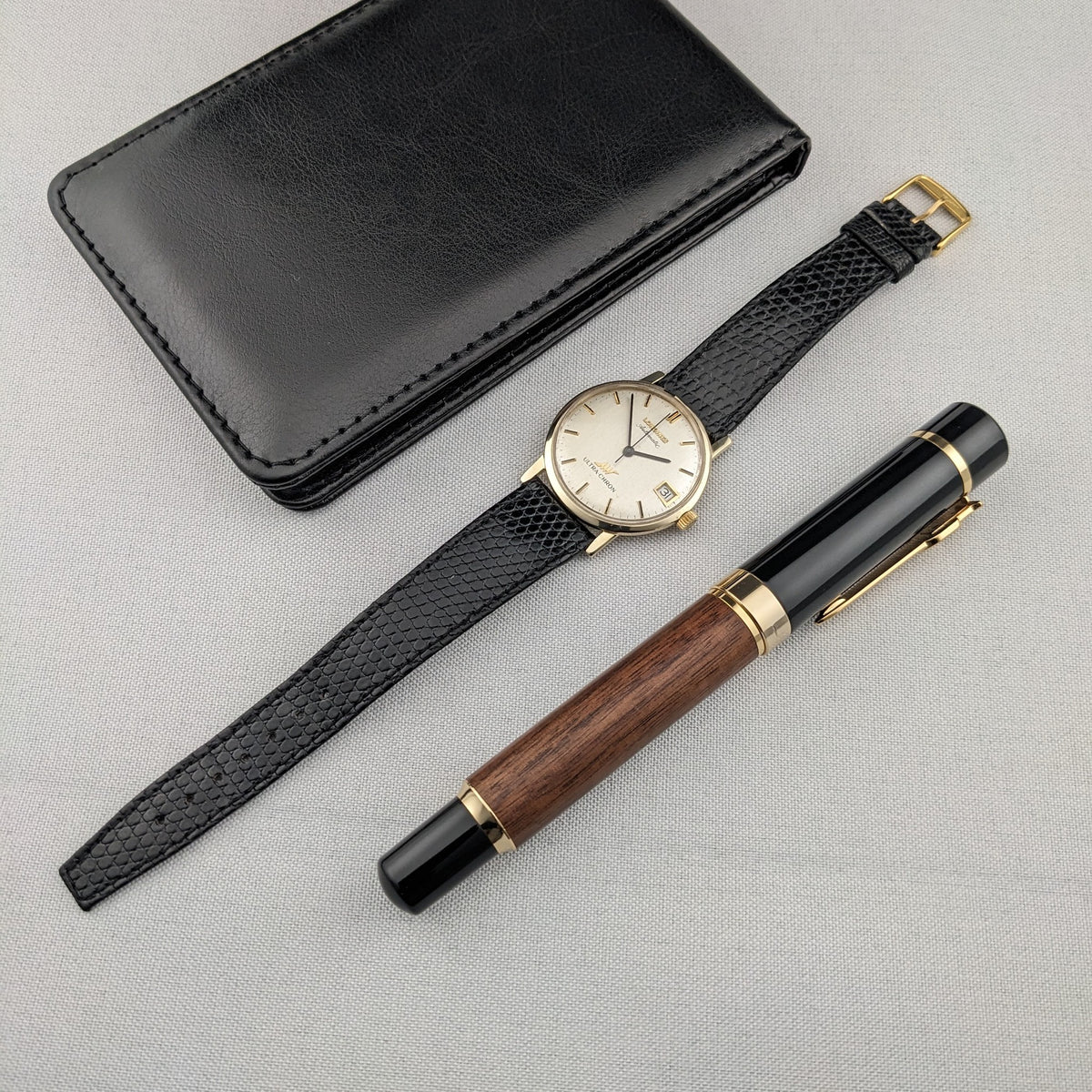 Full layout featuring the Longines Ultra-Chron 69-556AC, extended black strap, black case, and wood-and-black pen on light fabric.