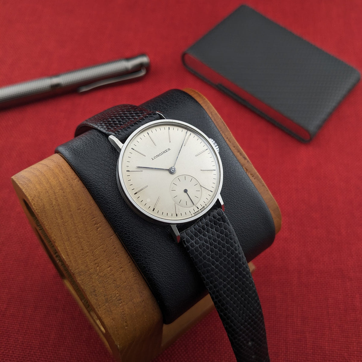 Longines Calatrava reference 20757 displayed on a black-and-wood watch cushion, with a notebook and silver pen behind it.