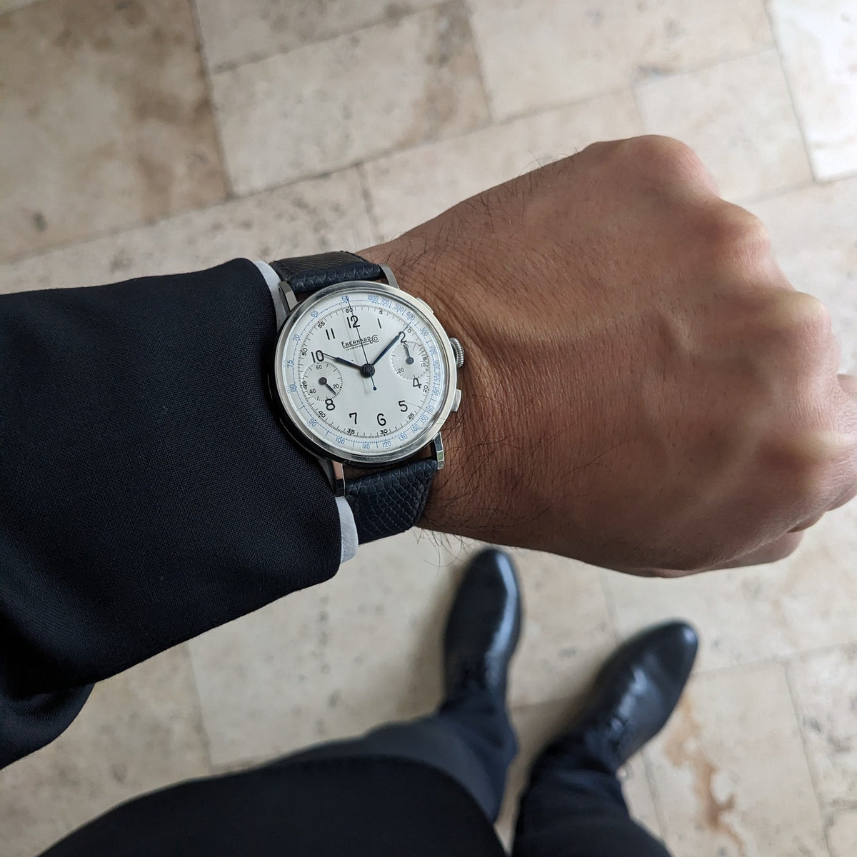 Eberhard & Co.. Pre-Extra Fort worn on a wrist with a dark blue strap, photographed above a tiled floor and polished shoes.