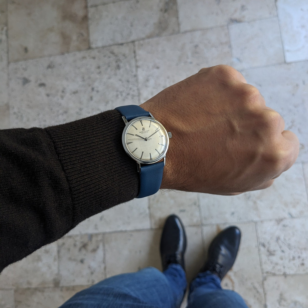 Breitling Automatic watch shown on a wrist from a wider viewpoint, with tiled flooring, jeans and black shoes visible below.
