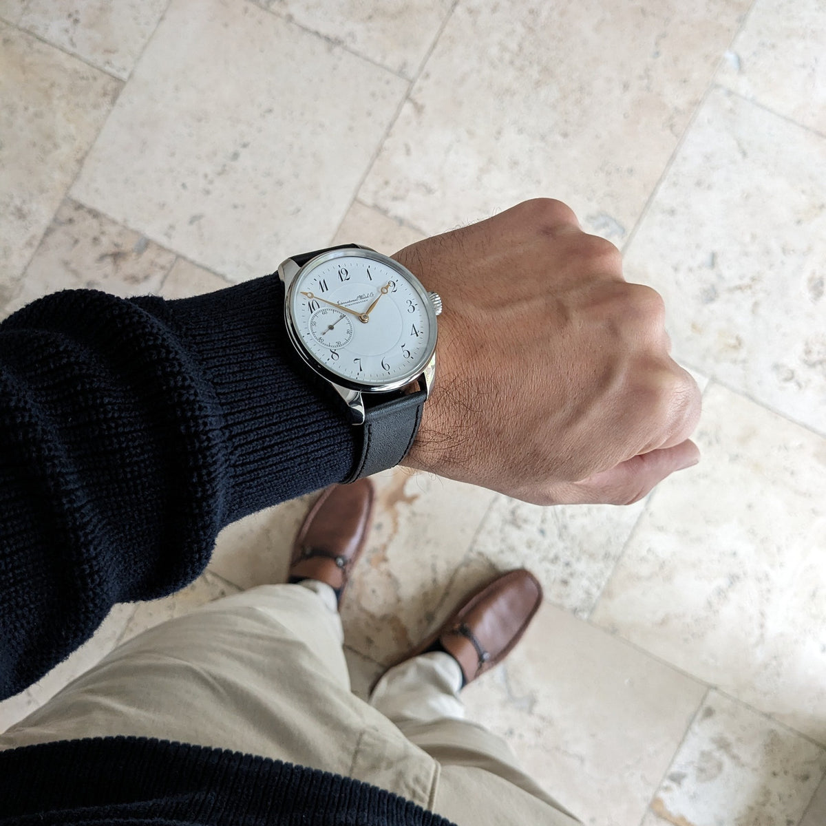 IWC Schaffhausen घड़ी का कलाई स्तर दृश्य, सफेद डायल और काले पट्टा के साथ, टाइल फर्श के खिलाफ दिखाया गया जिसमें पतलून और जूते दिखाई दे रहे हैं।