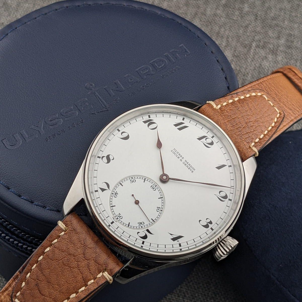 नीले ब्रांडेड केस पर Ulysse Nardin डायल का क्लोज़ व्यू, काले अंक, पतले हाथ और छोटे सेकंड सबडायल दिखाते हुए।