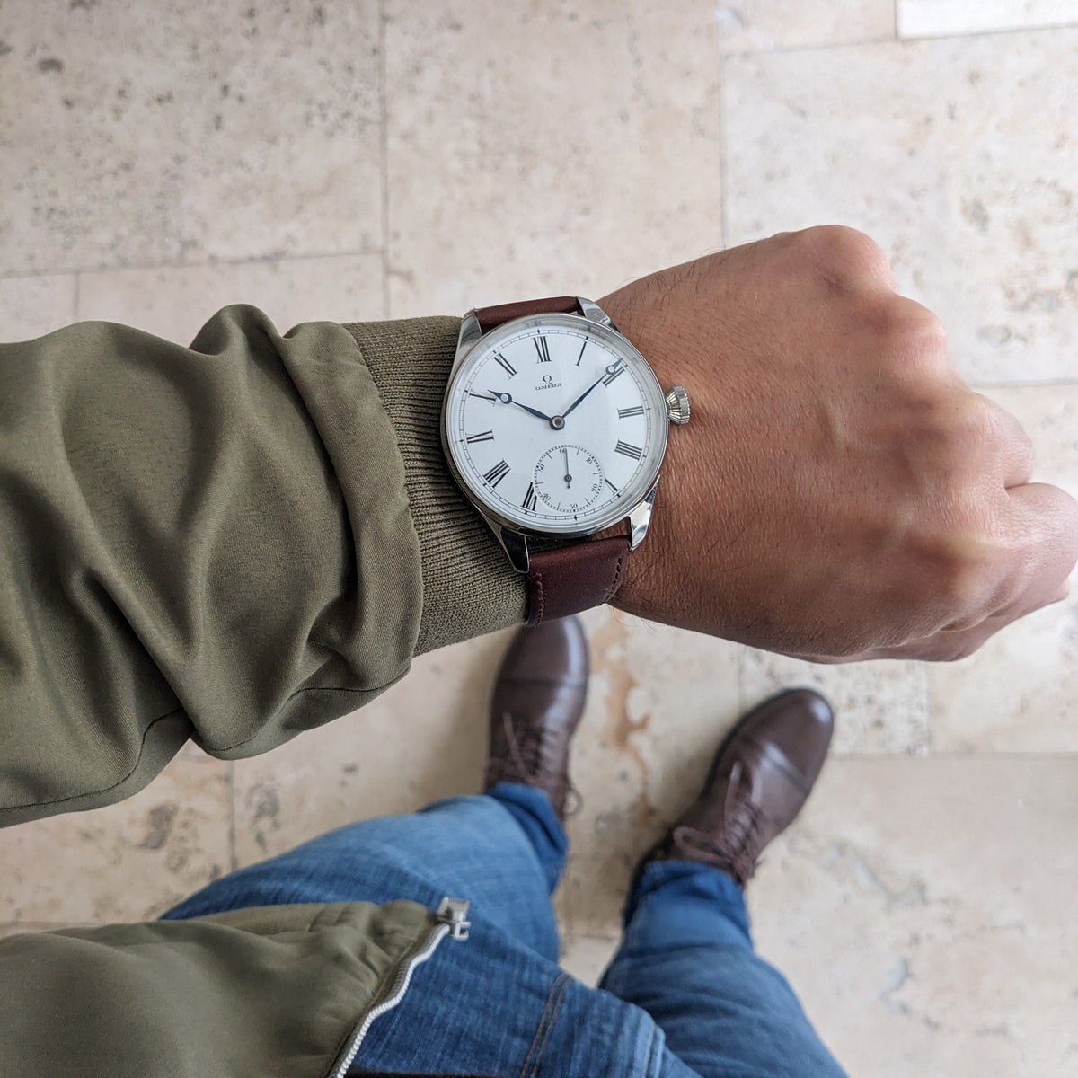 Vista più ampia di un orologio Omega con quadrante bianco e cinturino marrone, fotografato su pavimento piastrellato con abbigliamento e scarpe visibili.