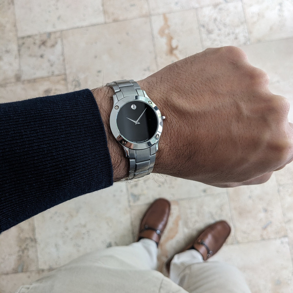 Vue plus large au poignet d'une montre Movado avec cadran noir et bracelet en acier, photographiée sur un sol carrelé.
