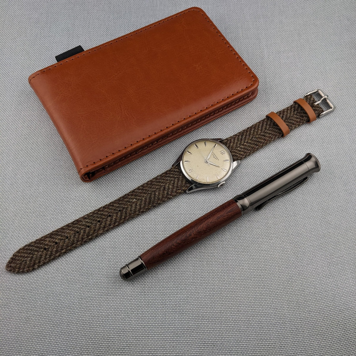 Longines Classic reference 8135-5 arranged with its brown woven strap, tan leather wallet and dark wood pen on gray fabric.