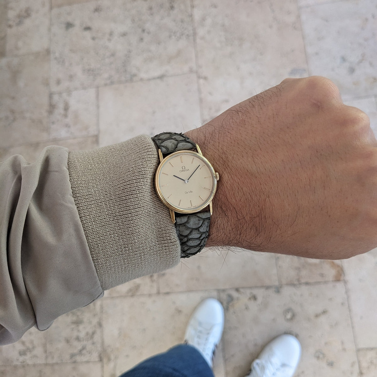 Wrist view of the Omega De Ville reference 111 0156 with cream dial and gray strap, photographed above a pale stone walkway.