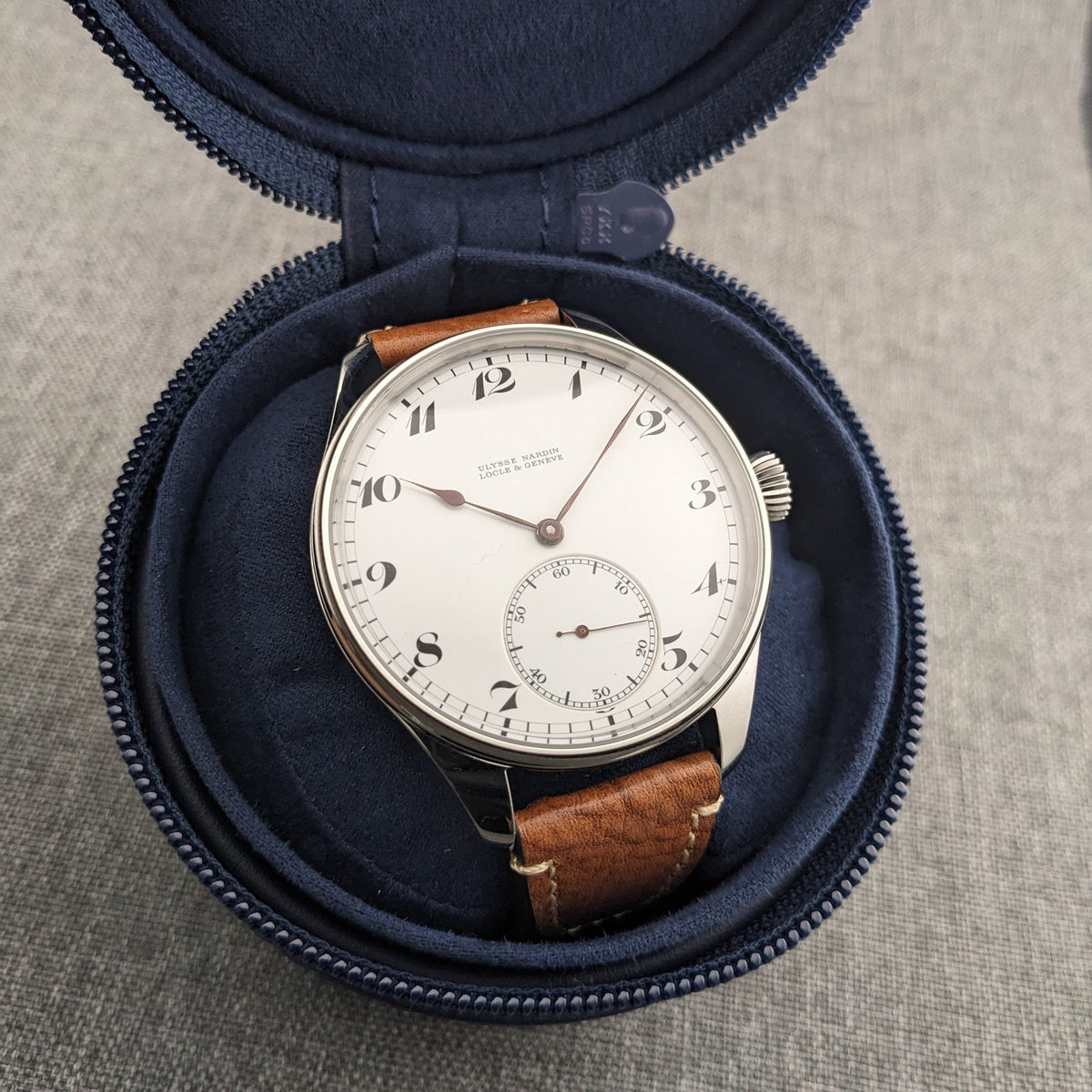 Ulysse Nardin कलाई घड़ी एक खुले नीले ज़िप केस के अंदर प्रदर्शित, जिसमें सफेद डायल और भूरा चमड़े का पट्टा पूरी तरह से दिखाई देता है।