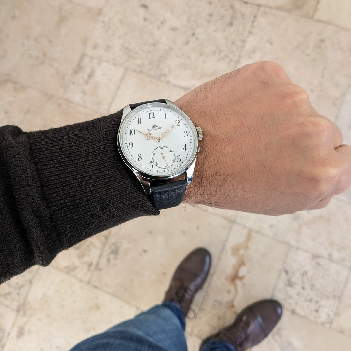 Deutsche Präzisionsuhr „Original Glashütte” am Handgelenk über einem Steinboden, mit vollem Zifferblatt und schwarzem Leder