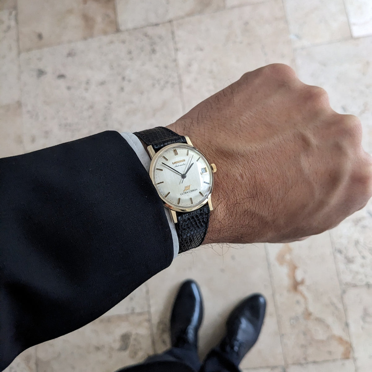 Wrist-level view of the Longines Ultra-Chron 69-556AC beneath a dark sleeve, with tiled flooring and black shoes in the background.