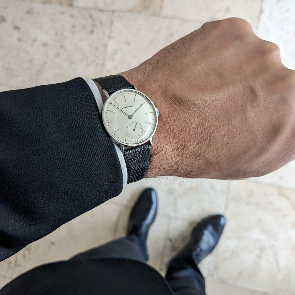 Longines Calatrava reference 20757 worn on a wrist with a dark suit sleeve, photographed above a pale floor with black shoes below.