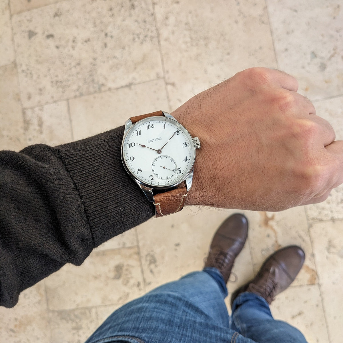 Ulysse Nardin कलाई घड़ी एक व्यक्ति की कलाई पर पत्थर के फर्श के ऊपर दिखाई गई, जिसमें भूरा पट्टा, सफेद डायल और छोटे सेकंड सबडायल दिखता है।