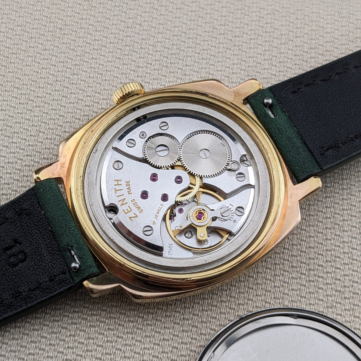 A close-up of a vintage Zenith watch showing its gold-tone case, transparent back, and intricate mechanical movement. The watch has a dark green leather strap and is photographed on a textured beige fabric background, with the detached silver metal back cover visible in the bottom right corner.
