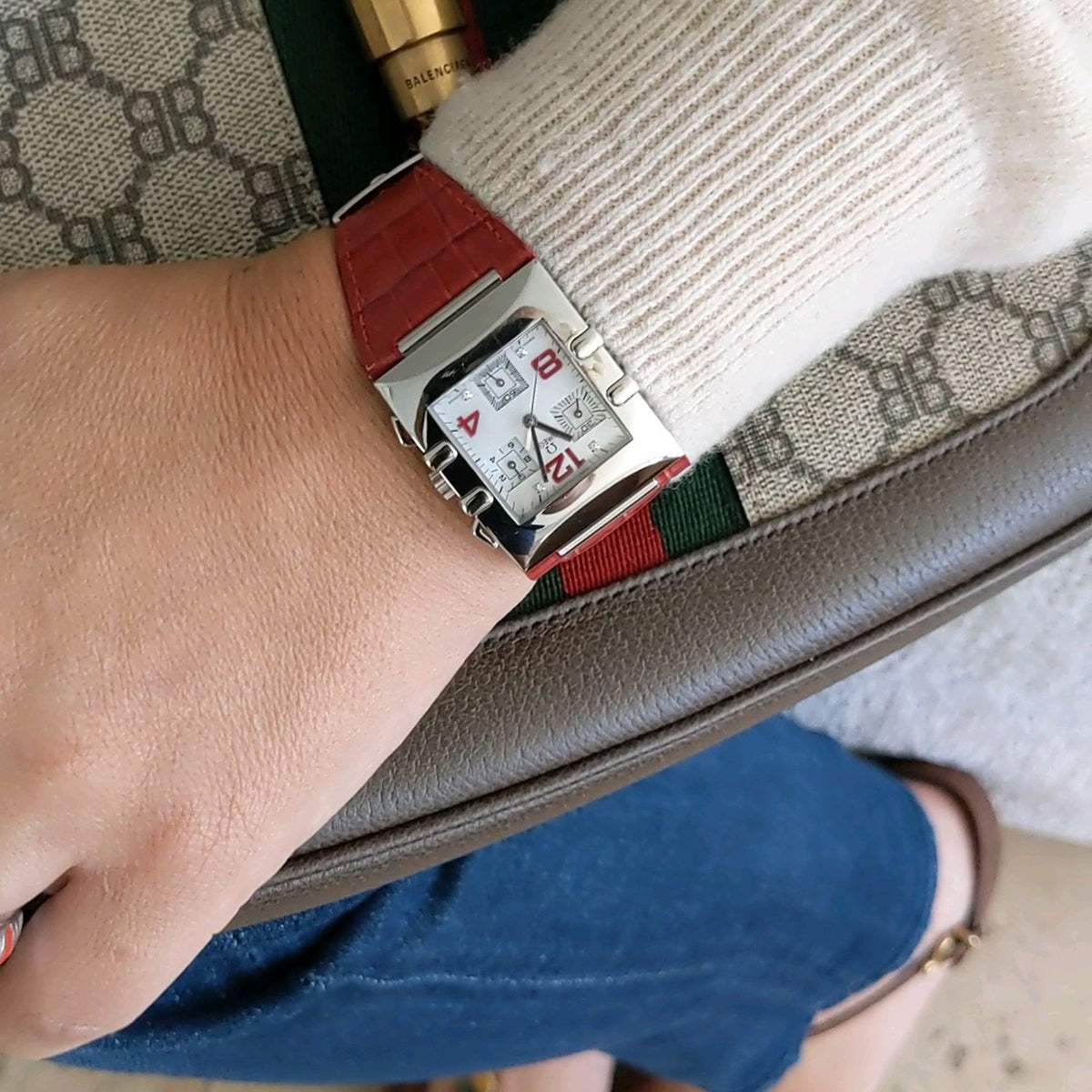 Omega Constellation Quadra 1841.73.31 worn on a wrist beside a patterned bag and blue clothing, with its red strap visible.
