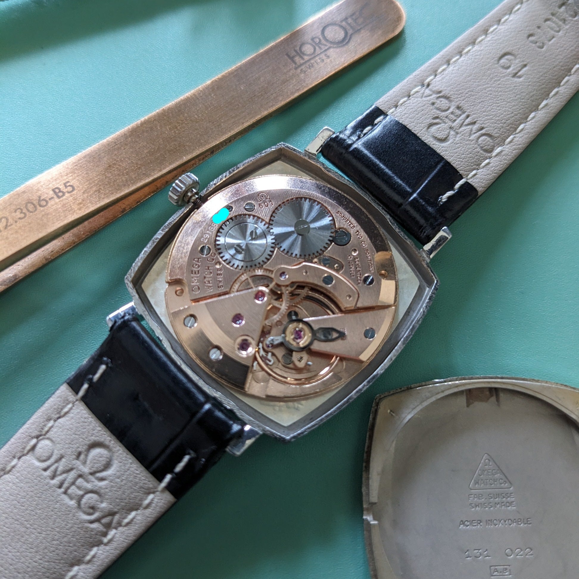 A close-up of an Omega automatic watch with its back cover removed, showing the intricate mechanical movement. The watch has a silver-toned case, a black leather strap, and a beige leather strap end.