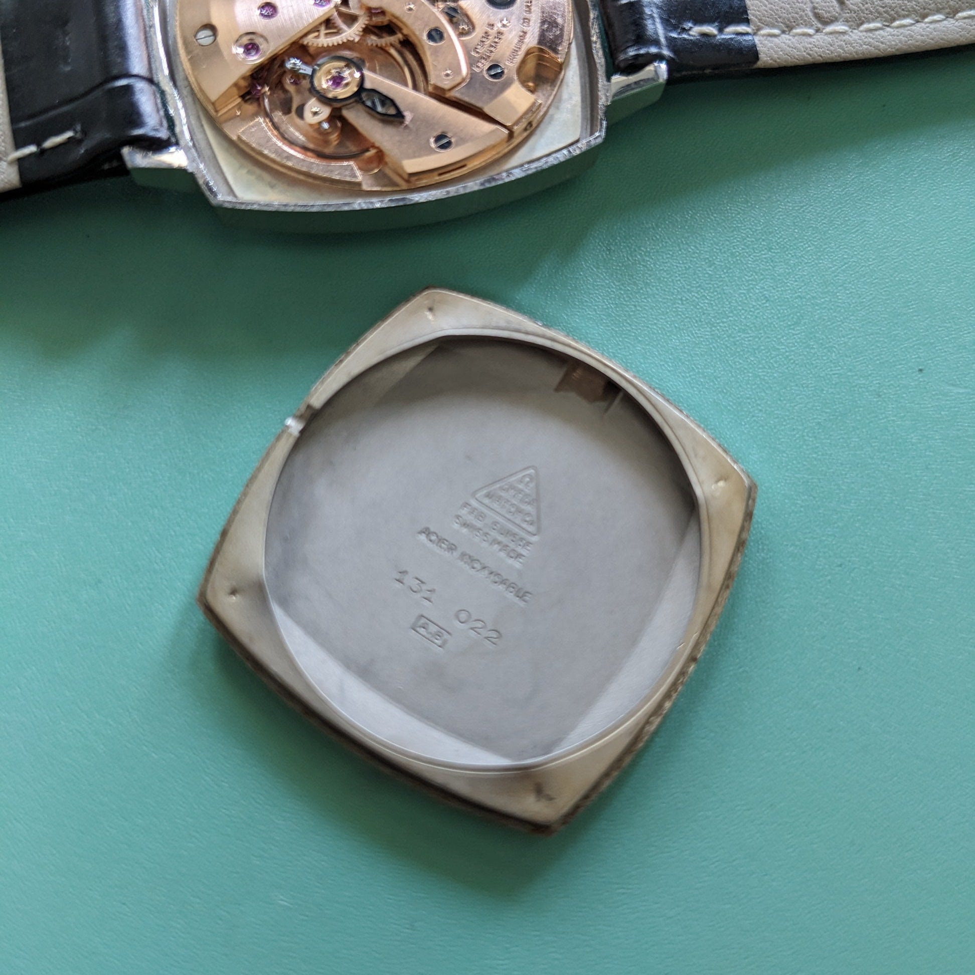 A close-up of a wristwatch showing the open back case with the exposed mechanical movement and a detached square-shaped back cover placed below it, on a teal-green background. The brand name 'OMEGA' is visible on the strap.