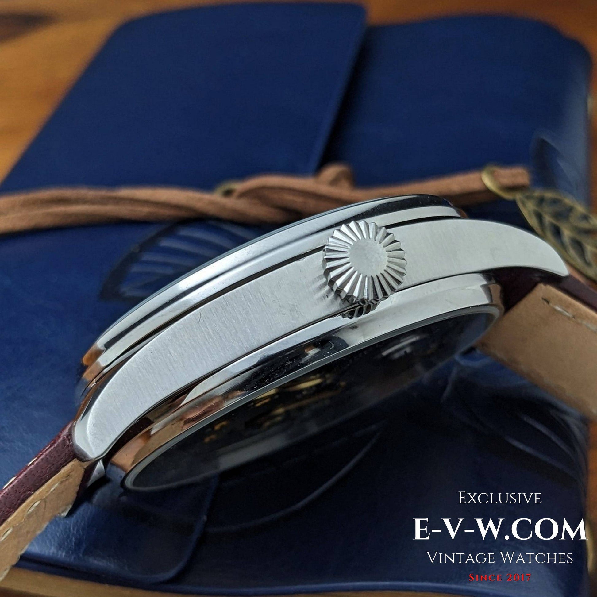 Close-up of a vintage watch with a brown strap on a blue background, featuring the brand 'E-V-W.com'.