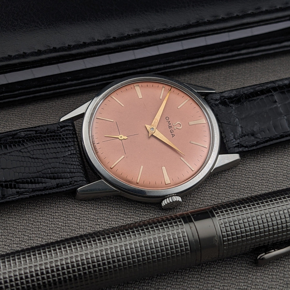 Un orologio Omega con quadrante rosa dorato e cassa argentata, su superficie grigia accanto a una penna nera e custodia in pelle nera.
