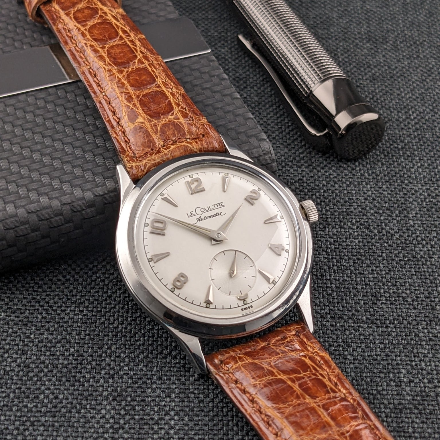 Jaeger-LeCoultre Automatic E328 lies across a black card holder beside a textured metal pen, showing its brown strap and dial.
